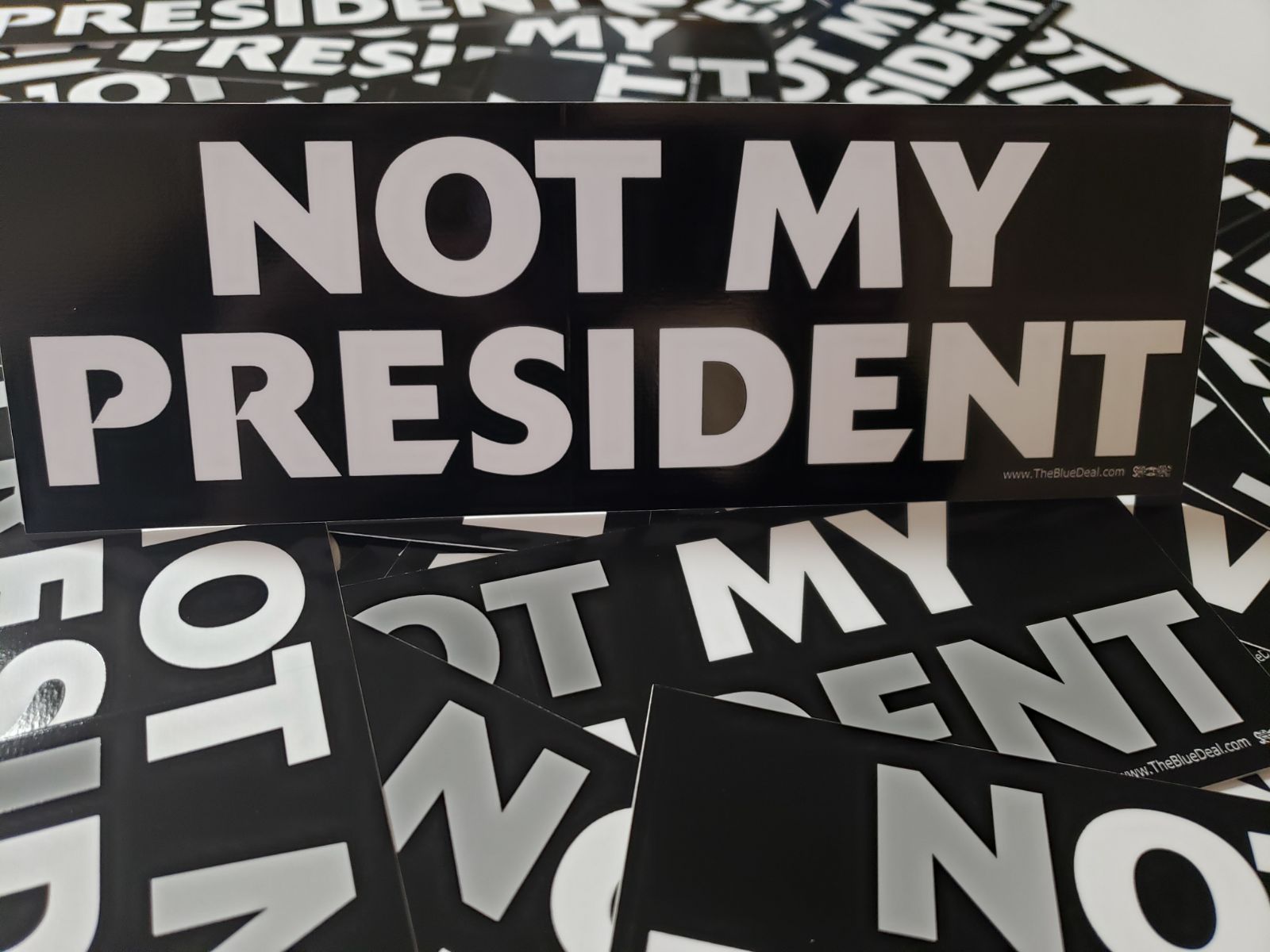 Close-up photo of a black bumper sticker with bold white text that reads ‘NOT MY PRESIDENT.’ The sticker is displayed on top of several identical stickers spread out beneath it.