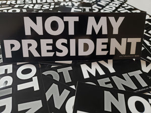 Close-up photo of a black bumper sticker with bold white text that reads ‘NOT MY PRESIDENT.’ The sticker is displayed on top of several identical stickers spread out beneath it.